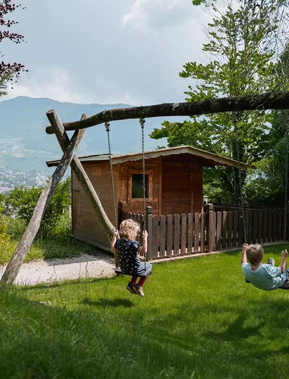 Zwei Kinder schaukeln vor einem kleinen Holzhaus